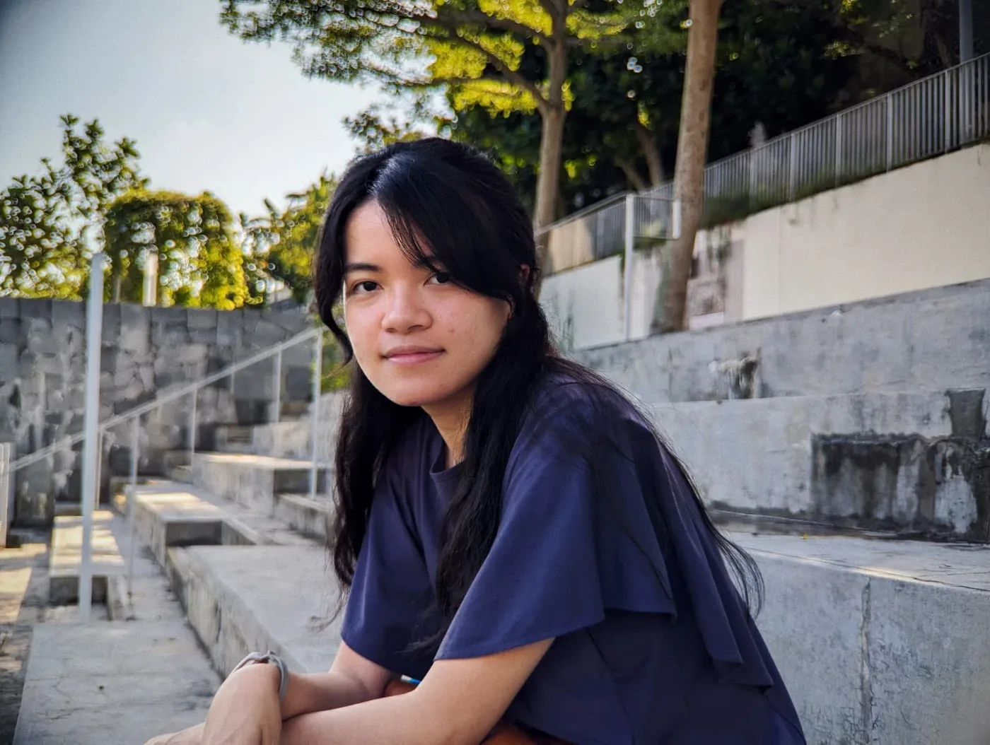 A young woman with long dark hair sitting on outdoor concrete steps, wearing a navy blue shirt, looking at the camera with trees and a fence in the background.