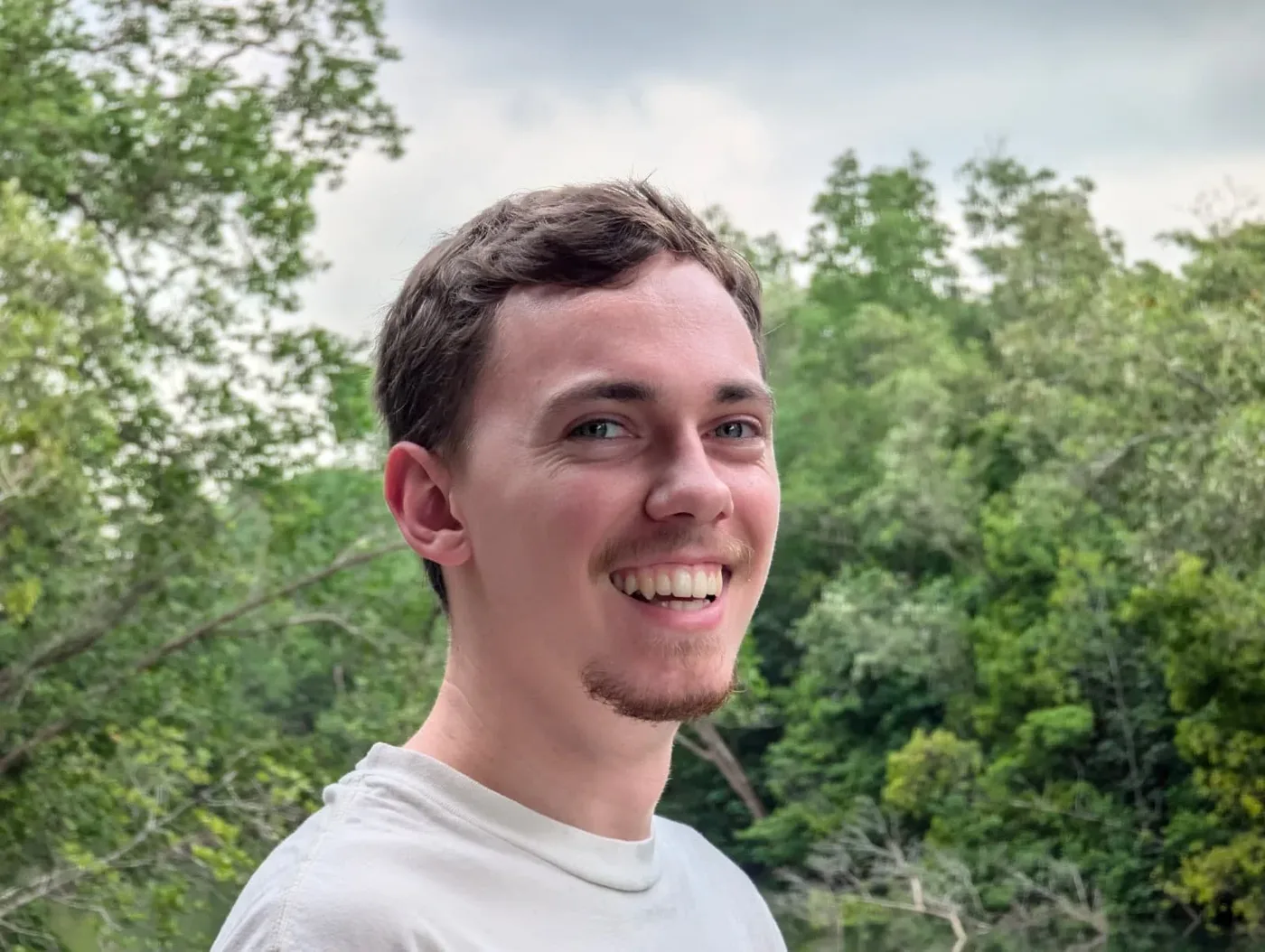 A young man with brown hair, blue eyes, and a goatee smiling outdoors with green trees and cloudy sky in the background.