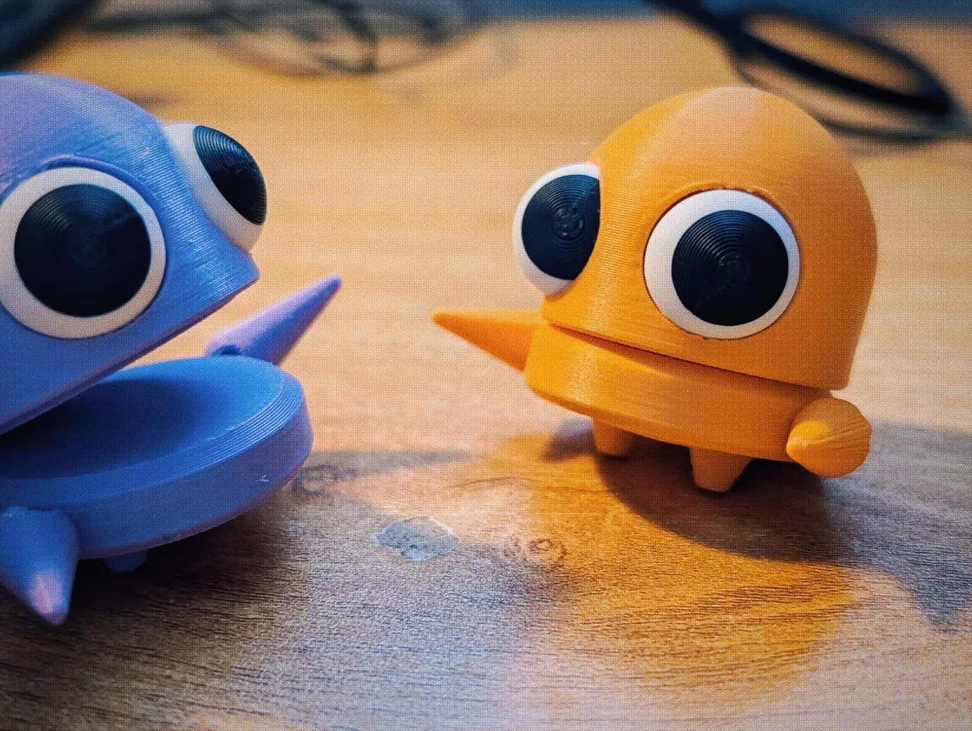 Colorful plastic toy creatures with large googly eyes on a wooden surface, one blue and one yellow.